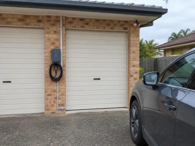 Wall-mounted EV charger with coiled cable on a brick garage wall beside double roller doors in Coombabah